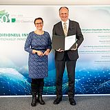 Sylvia Schattauer und Matthias von Kaler stehen gemeinsam vor der TU-Foto-Wand mit der aufgeklappten Urkunde.